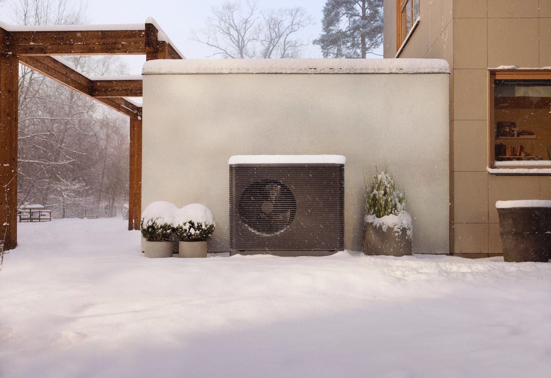 An Aira air source heat pump outdoor unit covered in snow with snow on the ground. It is placed against a wall, between some potted plants