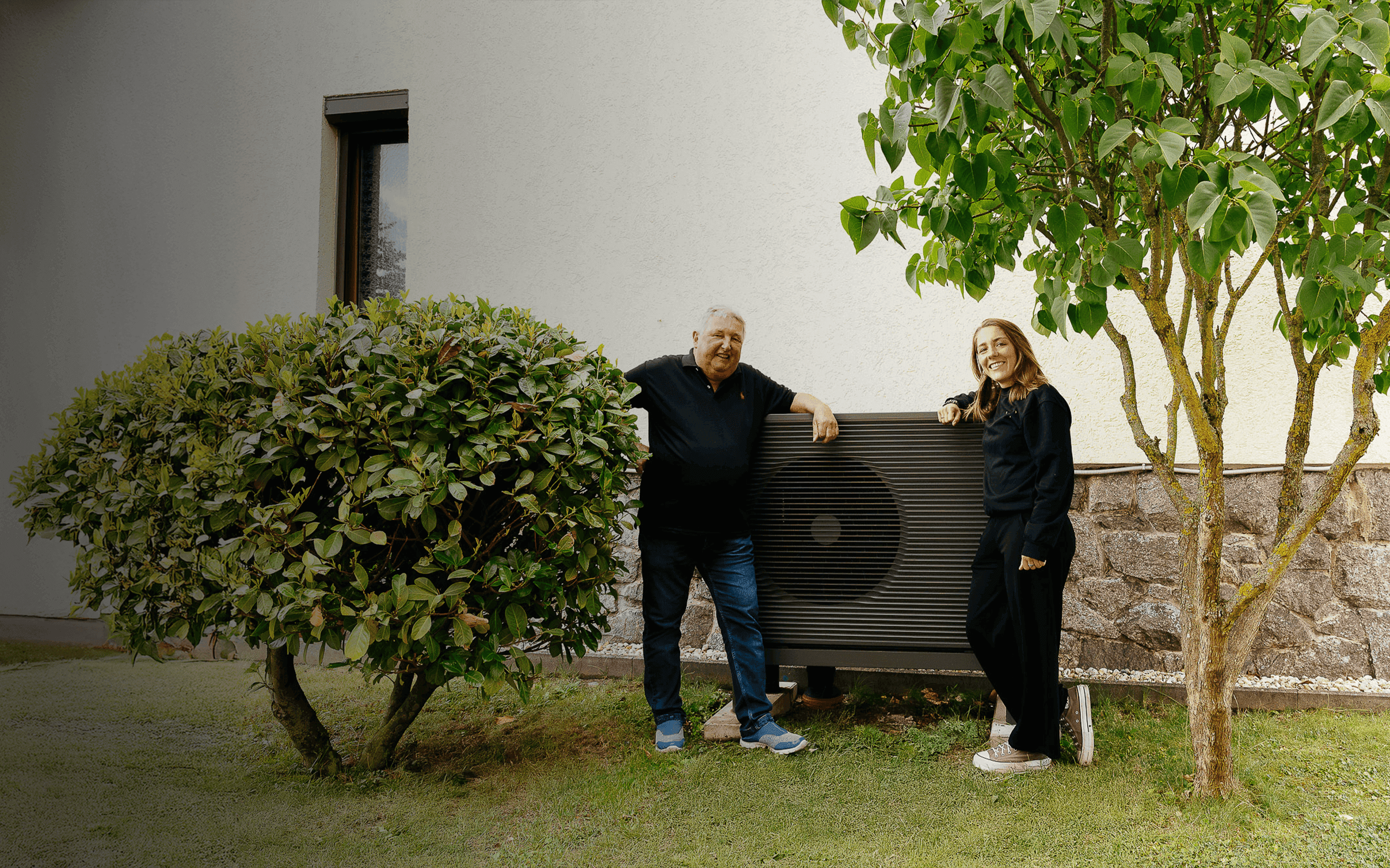 Älterer Mann und jüngere Frau stehen vor einer Wärmepumpe im Garten an einer weißen Hauswand